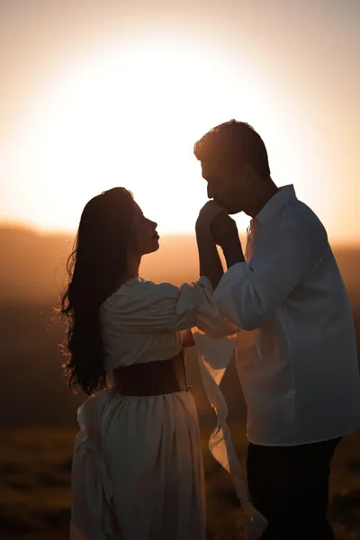 Joyce e Paulo Vinícius em silhueta contra o pôr do sol, ele beijando a mão dela